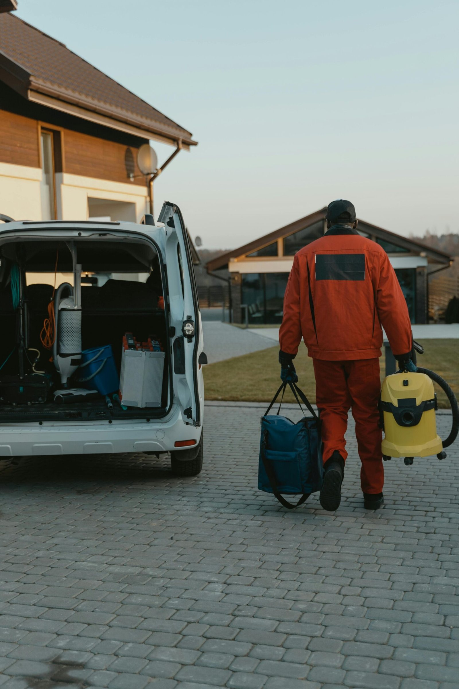 A worker in red work clothes carries a vacuum cleaner and bag near a van during the day.